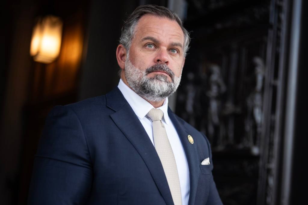 A man with a gray beard and short hair, wearing a navy suit and light-colored tie, stands in front of a dark, ornate background. He looks serious and professional.
