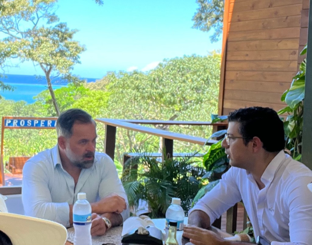 Two men engaged in conversation at a table outdoors, surrounded by lush greenery and an ocean view in the background.